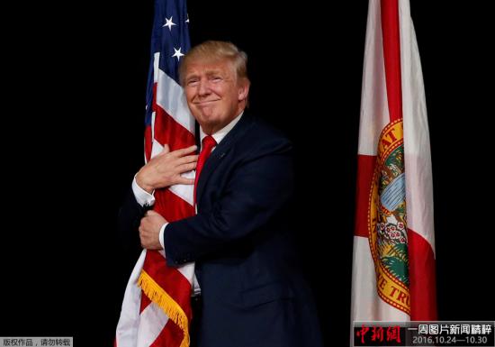 当地时间2016年10月24日，美国佛罗里达州坦帕市，美国共和党总统候选人唐纳德·特朗普在出席拉票活动时拥抱美国国旗。